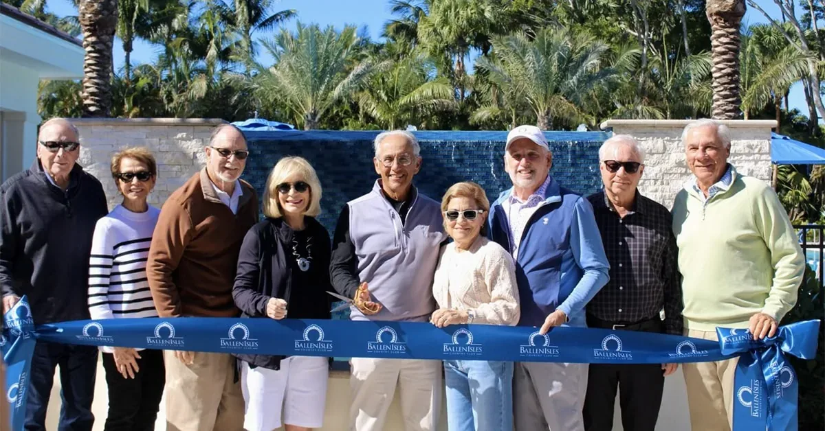A group of people participate in a ribbon-cutting ceremony at a luxury resort property with palm trees and a water feature in the background.