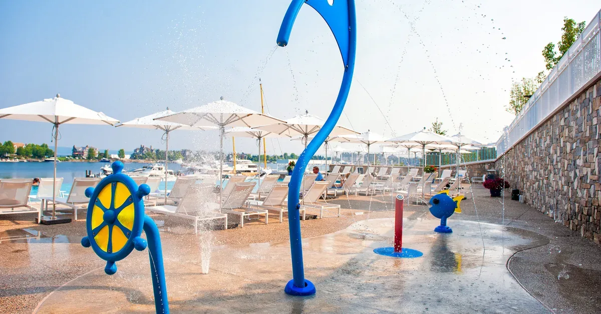 Waterfront resort features colorful children's splash pad with nautical-themed water features overlooking beach chairs and umbrellas by the lake.