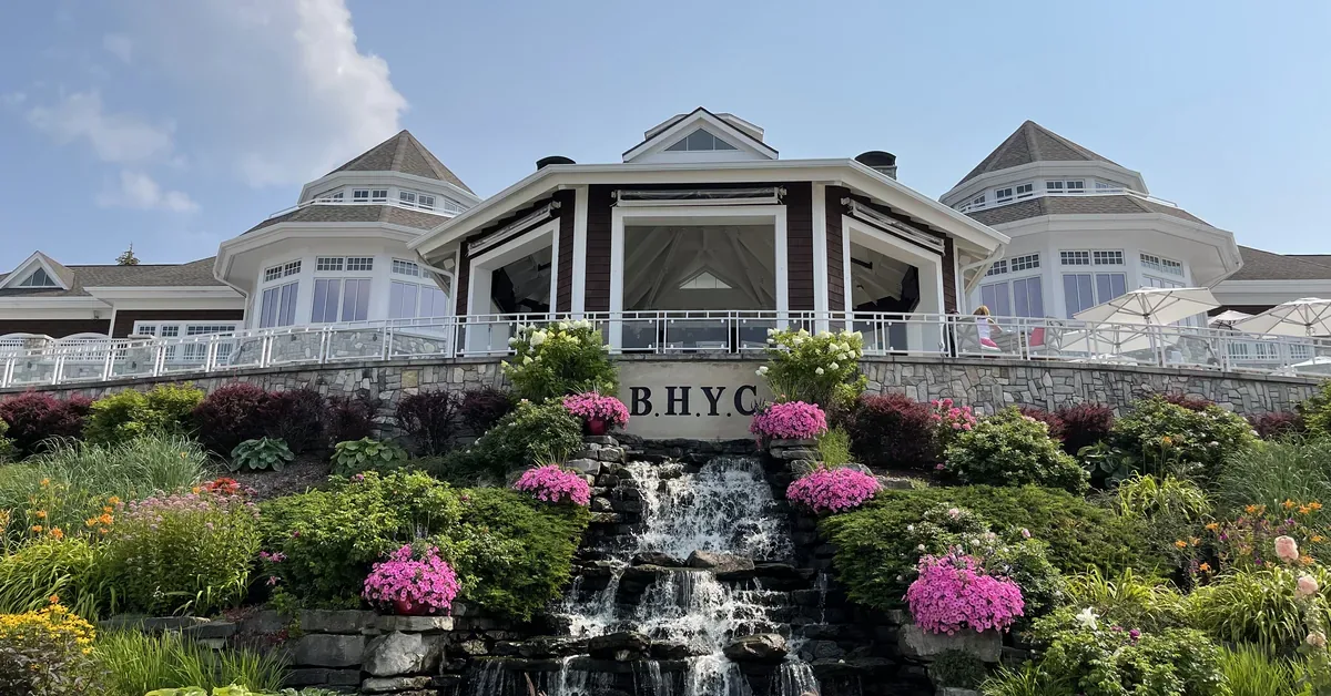 Elegant waterfront yacht club building features stone terracing, cascading waterfall, vibrant landscaping with purple flowers, and expansive windows overlook...