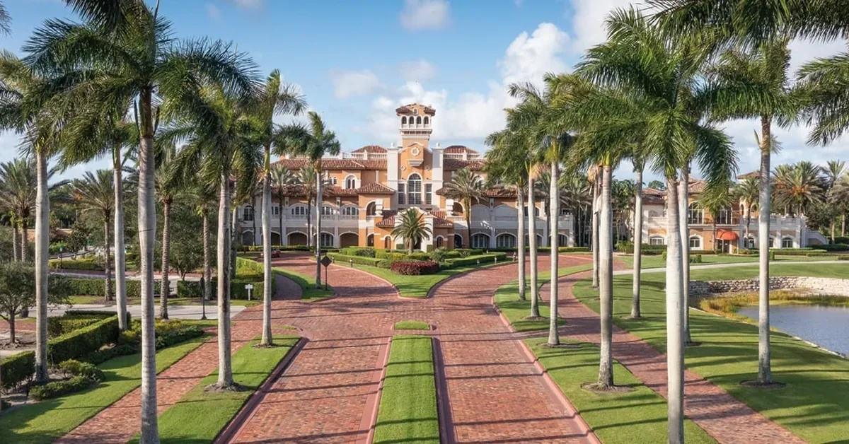 Luxurious Mediterranean-style mansion features an elegant clock tower and grand brick walkway lined with towering palm trees and manicured grounds.